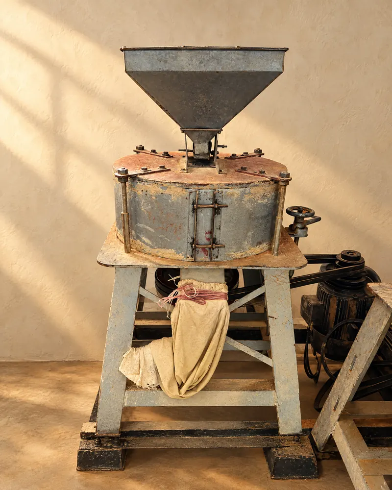 The flour mill in the SHRI JI FOODS unit: a steel hopper over a round mill housing on a steel stand, with a cloth outlet sleeve.